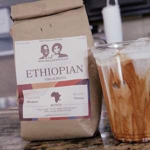 Puede incluir: Una bolsa de papel marrón con una etiqueta que dice "Ethiopian Yirgacheffe" de Hogans Coffee & Co. La etiqueta también incluye detalles sobre el origen del café, el proceso, el tueste y las notas de sabor. Un vaso de café helado está al lado de la bolsa.
