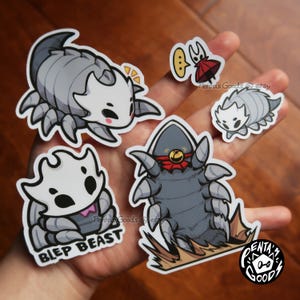 Puede incluir: Una colección de pegatinas de dibujos animados con varias criaturas grises y blancas con forma de insecto. Una pegatina dice "BLEP BEAST". Las pegatinas están en una mano, con un fondo de madera.