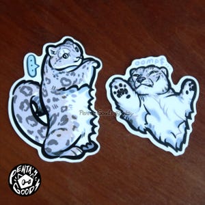 May include: Two stickers featuring cartoon snow leopards. One sticker shows a snow leopard with a thought bubble, the other shows a snow leopard with the word "oompf". The stickers are white with black outlines and a light purple and gray color scheme.