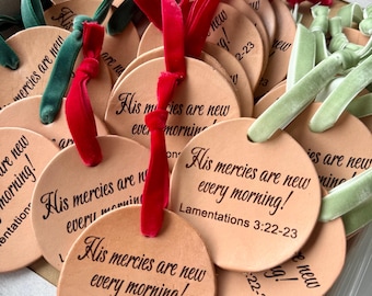 May include: A collection of round, tan leather ornaments featuring the black printed text 'His mercies are new every morning! Lamentations 3:22-23'. Each ornament is attached to a velvet ribbon in shades of red, dark green, or light green.