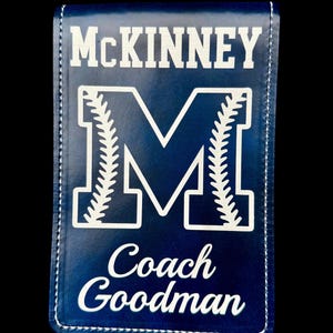May include: A navy blue baseball coach's notebook with white stitching. The notebook features the name "McKinney" at the top, a large white "M" with baseball stitching, and the name "Coach Goodman" at the bottom.