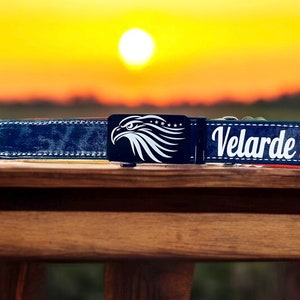 May include: A blue leather belt with a silver buckle featuring an eagle design. The belt has a red strap and the text "Velarde 14" is printed on the strap.