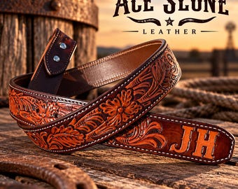 Può includere: Una cintura in pelle marrone con un intricato motivo floreale inciso, cuciture bianche a contrasto e le iniziali JH impresse sull'estremità. Il testo ACE SLONE LEATHER è visibile nell'angolo in alto a destra sopra la cintura.