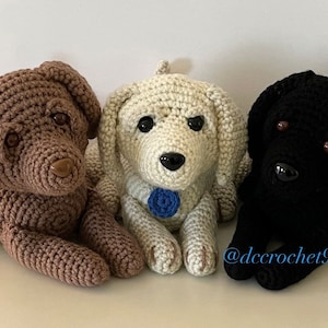 Op de afbeelding: Drie gehaakte knuffels, een bruine, een witte met een blauwe strik en een zwarte. De honden zitten naast elkaar op een wit oppervlak. De onderschrift '@dcccrochet9' is zichtbaar in de rechterbenedenhoek van de afbeelding.