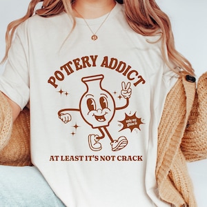 Może przedstawiać: Biała koszulka z brązowym nadrukiem przedstawiającym kreskówkowy wazon ceramiczny z tekstem "Pottery Addict" i "At least it's not crack".