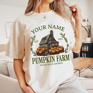 Op de afbeelding: Wit T-shirt met een afbeelding van een huis met pompoenen en de tekst "Your Name Pumpkin Farm Est 1962 Magically Grown".