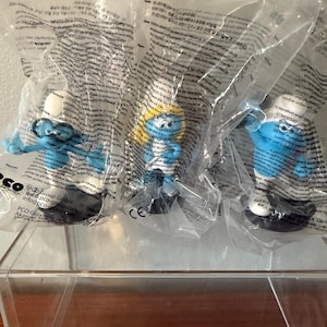 Może przedstawiać: Trzy figurki Smerfów w przezroczystym plastikowym opakowaniu. Smerfy są niebieskie z białymi czapkami i czarnymi butami. Jeden Smerf ma okulary, a drugi żółtą czapkę. Opakowanie ma nadrukowany tekst.