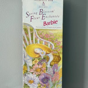 Peut inclure: Boîte de poupée Barbie Spring Blossom. La boîte présente le texte "Spring Blossom Fleur Enchantée Barbie" avec un motif floral et un chapeau jaune sur une chaise blanche. Il s'agit d'une édition spéciale d'Avon.