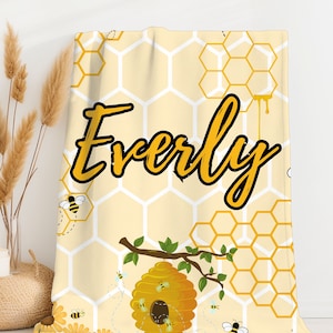 Puede incluir: Una manta amarilla y blanca con un patrón de panal y el nombre "Everly" escrito en negro. La manta presenta una colmena con abejas y una rama con hojas verdes.