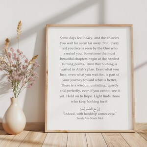 May include: A framed wall art print featuring black text on a white background. The text reads: Some days feel heavy, and the answers you wait for seem far away. It also includes the Arabic script and the English translation: Indeed, with hardship comes ease. Surah Ash-Sharh 94:6. A beige vase with dried flowers sits beside the frame.