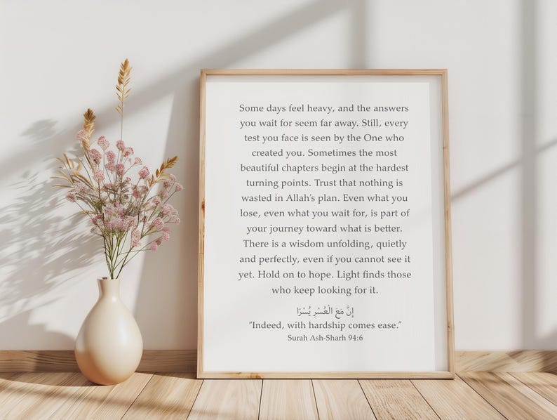 May include: A framed wall art print featuring black text on a white background. The text reads: Some days feel heavy, and the answers you wait for seem far away. It also includes the Arabic script and the English translation: Indeed, with hardship comes ease. Surah Ash-Sharh 94:6. A beige vase with dried flowers sits beside the frame.