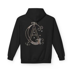 Peut inclure: Sweat à capuche noir avec un motif de monogramme doré au dos. Le monogramme comprend les lettres "A" et "C" entrelacées avec des ornements décoratifs et des étoiles. Le motif est centré au dos du sweat à capuche.