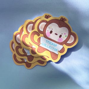May include: A pile of stickers depicting a cartoon monkey. The monkey has a light brown face, ears, and limbs, and a yellow body. It wears a white shirt with the text "STICKER COLLECTOR". The monkey has pink cheeks and colourful accents.