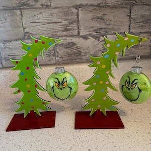 May include: Two Christmas tree ornaments with a Grinch face design. Each ornament is a green glass ball with a Grinch face. The ornaments hang from a silver hook attached to a green wooden Christmas tree. The trees are on a red base.