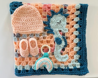 May include: A crochet set featuring a peach-colored beanie, a pair of matching peach booties, a light blue seahorse applique, and a wooden ring rattle with a small rainbow attachment, all resting on a peach and teal square crochet blanket.