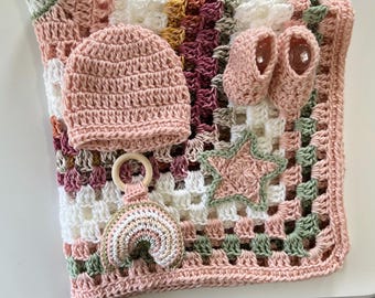 May include: A crochet baby set featuring a multi-colored granny square blanket in shades of pink, white, and sage green. The set includes a solid pink beanie, a pair of matching pink booties, a wooden ring rattle, and a small crocheted rainbow.