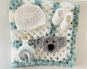 May include: A handmade baby set featuring a crocheted blanket with a koala applique, a white beanie, and a pair of baby booties. A rainbow-shaped teether is also included. The blanket has a white, blue, and green color scheme.