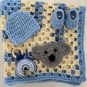 May include: A crochet baby set featuring a cream and blue granny square blanket, a light blue beanie, a pair of light blue booties, a gray koala face appliqué, and a wooden ring toy with a blue and white crocheted rainbow attachment.