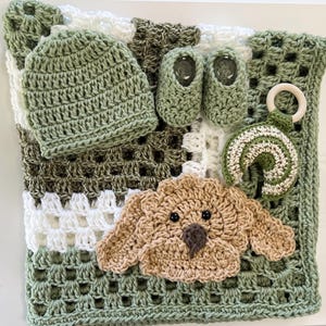 May include: A sage green crochet blanket with a granny square pattern in white and olive green. Included are a matching sage green beanie, a pair of sage green booties, a rainbow-shaped rattle with a wooden ring, and a tan crochet dog applique.