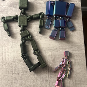 May include: Three articulated robot figures in varying sizes. The largest is dark green, the medium-sized one is iridescent blue, and the smallest is iridescent pink. Each figure has a block-shaped head and body, with jointed limbs.