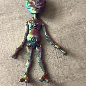 May include: A colorful alien figurine with a metallic, iridescent finish. The figure has a large head with prominent eyes and small antennae. The body is segmented, with articulated arms and legs. The alien is posed with arms outstretched, showcasing its unique design.