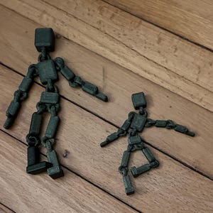 May include: Two articulated wooden figures, one large and one small, stand on a wooden surface. The figures are dark green and have a block-like head and limbs connected by joints, allowing for posing.
