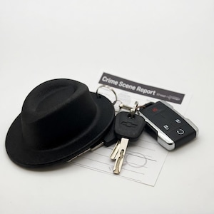 Può includere: Un cappello fedora nero, chiavi dell'auto e un portachiavi sono appoggiati su un documento con la scritta "Crime Scene Report". Le chiavi includono una chiave Chevrolet e un portachiavi per auto con dettagli argentati. Su sfondo bianco.