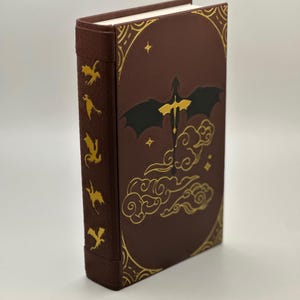 Puede incluir: Un diario encuadernado en cuero burdeos con un diseño de dragón dorado en la portada. El dragón está volando en las nubes con estrellas en el fondo. El lomo del diario tiene un diseño dorado de dragones volando.