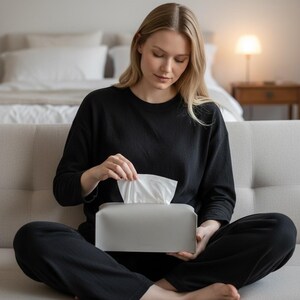 Puede incluir: Una persona vestida de negro, sentada en un sof&aacute; claro, saca un pa&ntilde;uelo de una caja de pa&ntilde;uelos gris. La caja es rectangular y de dise&ntilde;o minimalista. En el fondo, una cama y una l&aacute;mpara.