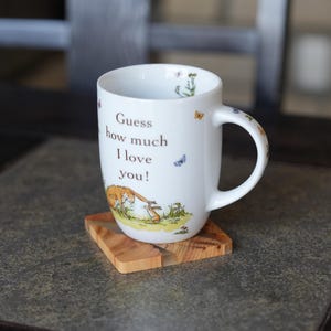 K&ouml;nnte beinhalten: Wei&szlig;e Keramiktasse mit dem Text "Guess how much I love you!" und Illustrationen eines Hasen und Schmetterlingen. Die Tasse steht auf einem kleinen, quadratischen Holzuntersetzer. Die Tasse hat einen gebogenen Griff.