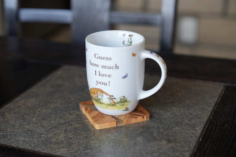 K&ouml;nnte beinhalten: Wei&szlig;e Keramiktasse mit dem Text "Guess how much I love you!" und Illustrationen eines Hasen und Schmetterlingen. Die Tasse steht auf einem kleinen, quadratischen Holzuntersetzer. Die Tasse hat einen gebogenen Griff.