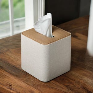 May include: A white tissue box cover with a light brown leather top and a white fabric base. The cover is sitting on a wooden surface.