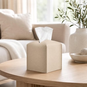May include: A square tissue box cover with a textured, light beige faux leather finish. The cover features a central rectangular opening at the top, through which a single white facial tissue is pulled upward, standing upright in a vertical position.