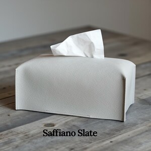A rectangular, light gray tissue box cover with a textured surface. A white tissue is pulled from the top opening. The cover has a simple, modern design, suitable for home or office use. This is purely made with designer quality Genuine Leather.