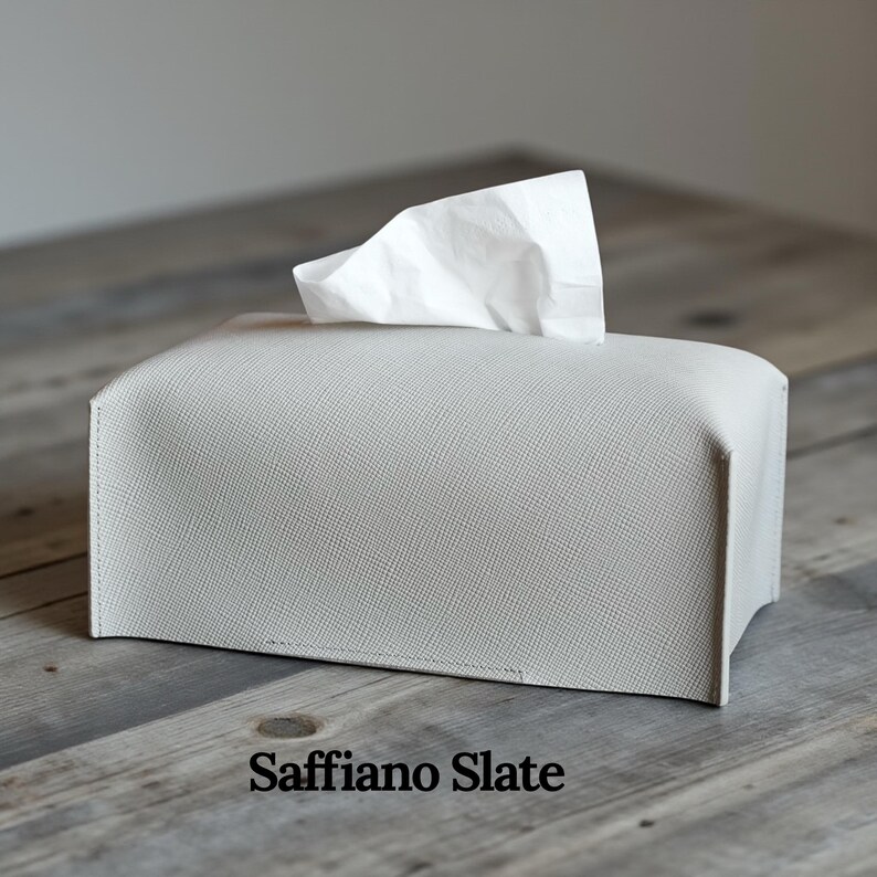 A rectangular, light gray tissue box cover with a textured surface. A white tissue is pulled from the top opening. The cover has a simple, modern design, suitable for home or office use. This is purely made with designer quality Genuine Leather.
