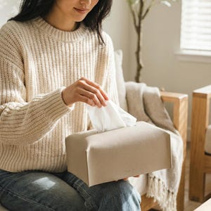 Puede incluir: Un dispensador de pa&ntilde;uelos rectangular beige con una superficie texturizada. Se ven pa&ntilde;uelos blancos en la parte superior. La caja la sostiene una persona que lleva un su&eacute;ter color crema y vaqueros azules.