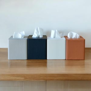 May include: Four square tissue box holders in a row, each with a different color and texture. The colors are light gray, navy blue, white, and orange-brown. Each box has a vertical ribbed design and holds a stack of white tissues.
