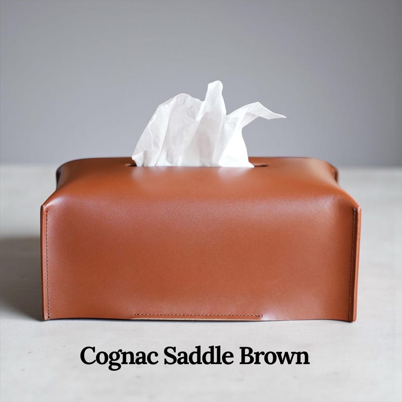 A rectangular, light gray tissue box cover with a textured surface. A white tissue is pulled from the top opening. The cover has a simple, modern design, suitable for home or office use. This is purely made with designer quality Genuine Leather.