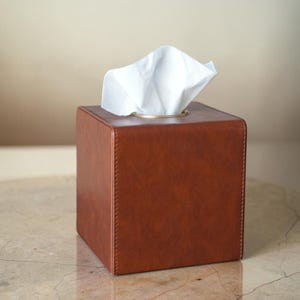 以下が含まれることがあります： 温かみのあるリッチなブラウンカラーの正方形のティッシュボックスカバーで、垂直の縁に沿ってステッチが見えます。ボックスの上部にはゴールドトーンの金属製リムが付いた円形の開口部があり、そこから白いティッシュが取り出せるようになっています。