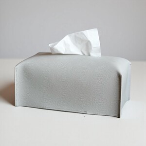 A rectangular, light gray tissue box cover with a textured surface. A white tissue is pulled from the top opening. The cover has a simple, modern design, suitable for home or office use. This is purely made with designer quality Genuine Leather.
