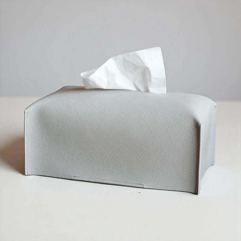 A rectangular, light gray tissue box cover with a textured surface. A white tissue is pulled from the top opening. The cover has a simple, modern design, suitable for home or office use. This is purely made with designer quality Genuine Leather.