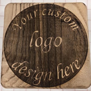 May include: A square wooden block with a circular, darker wood inlay. The inlay features the words "Your custom logo design here" carved into the wood. The wood grain is visible throughout the block and inlay.