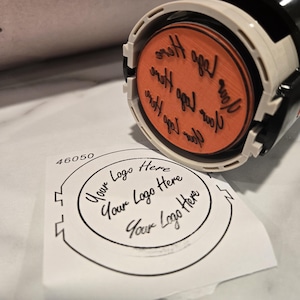 May include: A close-up of a self-inking stamp with a round orange ink pad. The ink pad has the text "Your Logo Here" written on it in black cursive. The stamp is on a white piece of paper with the same text printed in black.