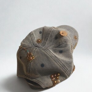 May include: A denim baseball cap with decorative embellishments. The cap is a faded brown color and features various beaded and pearl accents, along with a wooden flower. The cap has a curved brim and a classic six-panel design.