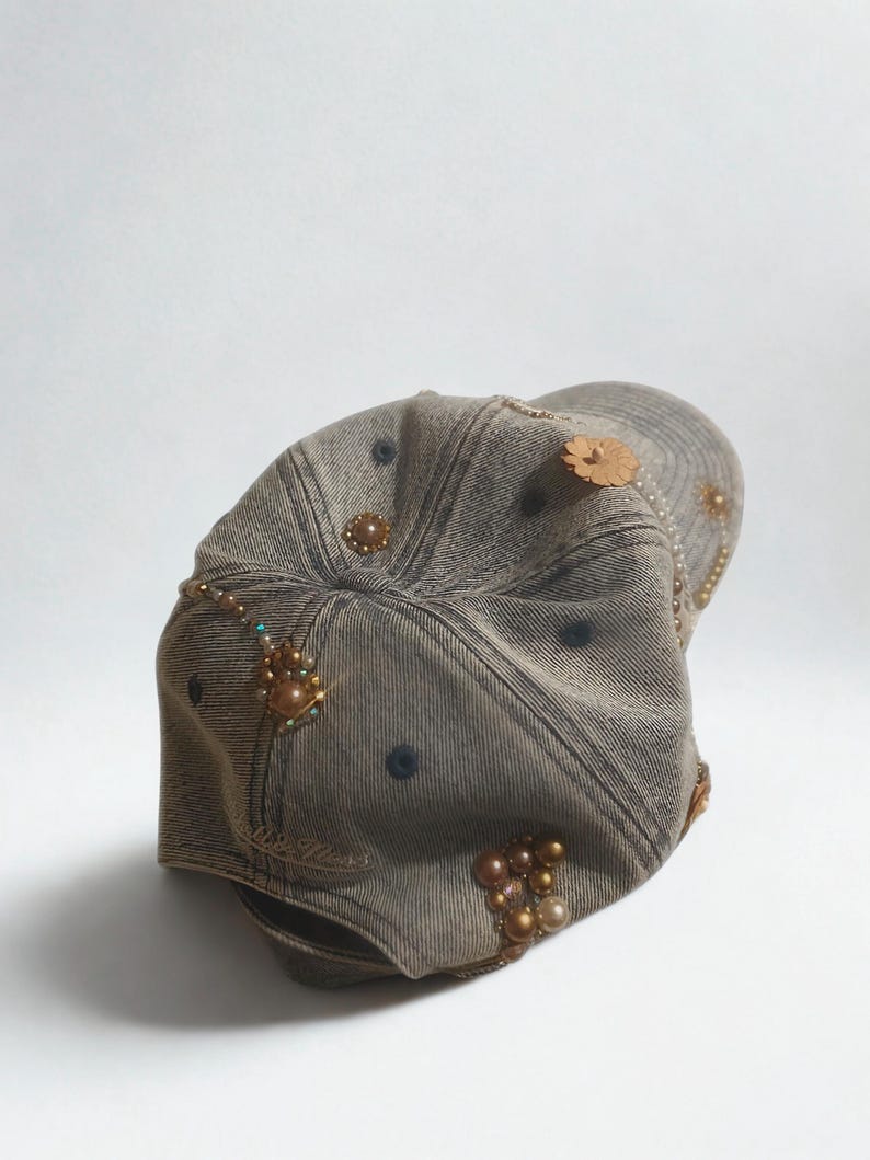May include: A denim baseball cap with decorative embellishments. The cap is a faded brown color and features various beaded and pearl accents, along with a wooden flower. The cap has a curved brim and a classic six-panel design.