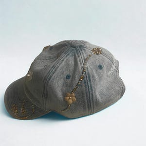 May include: Gray denim baseball cap with floral and beaded embellishments. The cap features a curved brim and a decorative design of small flowers and beads. The cap has a faded appearance and a curved brim. The brand name "Mitchell" is visible on the side.
