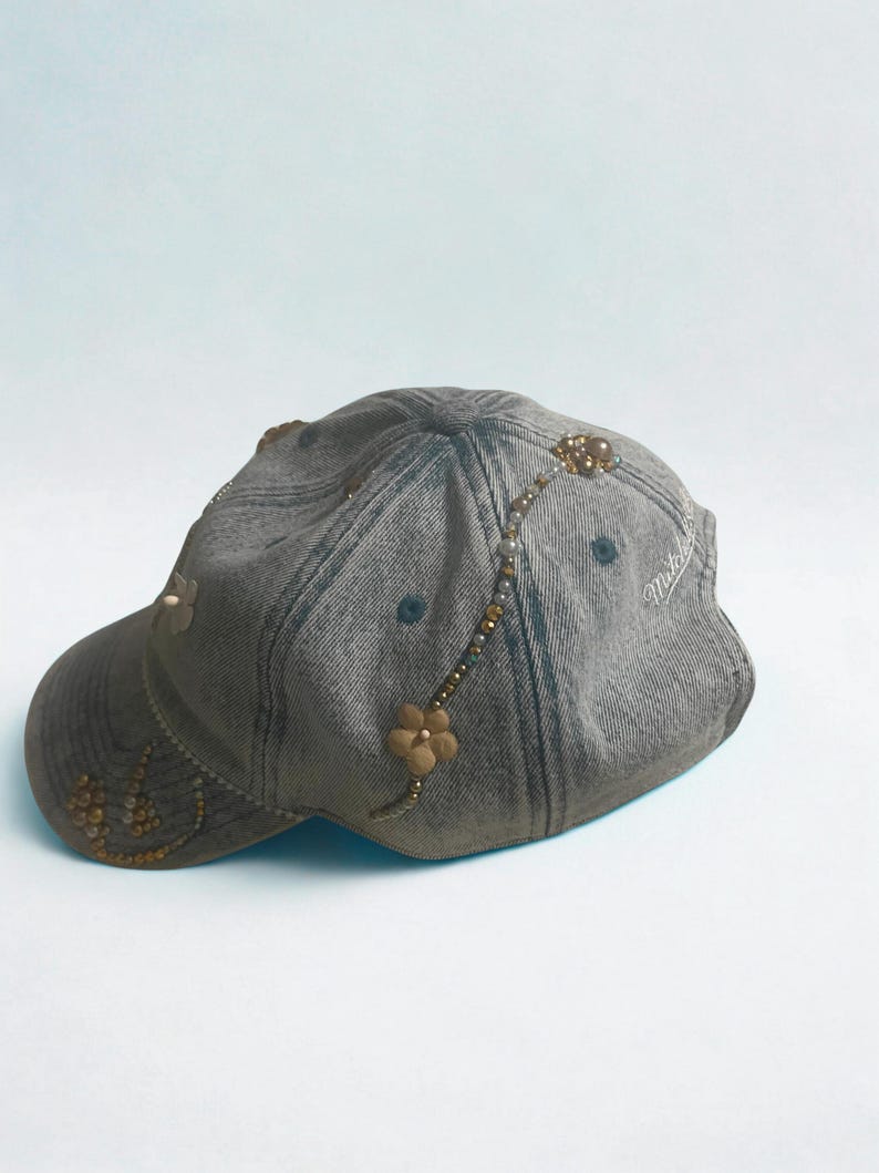 May include: Gray denim baseball cap with floral and beaded embellishments. The cap features a curved brim and a decorative design of small flowers and beads. The cap has a faded appearance and a curved brim. The brand name "Mitchell" is visible on the side.