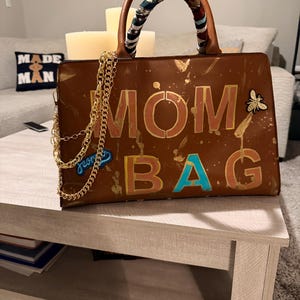 May include: Brown handbag with gold accents and the words "MOM BAG" in gold and turquoise. The bag has a gold chain strap and a patterned scarf wrapped around the handles. A small bee-shaped pin is attached.