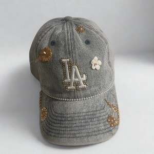 May include: Gray denim baseball cap with the letters "LA" in white, embellished with small beads. The cap features decorative floral accents in shades of brown and white, adding a touch of elegance to the casual design.