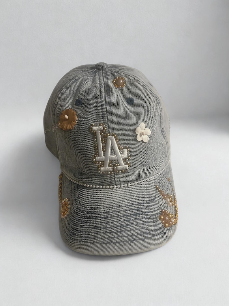 May include: Gray denim baseball cap with the letters "LA" in white, embellished with small beads. The cap features decorative floral accents in shades of brown and white, adding a touch of elegance to the casual design.